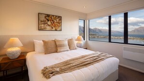 A bedroom with a large bed, two bedside lamps, a view of mountains and a lake, and a painting on the wall.