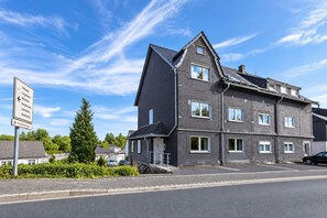 En flereetagers boligbygning med et stejlt tag, en tydelig indgang og et skilt, der viser vejen til nærliggende byer.