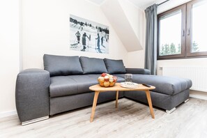 A modern living room with a grey sofa, a wooden coffee table with a fruit bowl and a glass, and a window with curtains.