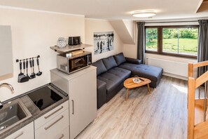 Cocina compacta con microondas, hervidor, utensilios, sofá, una pequeña mesa y ventana con vistas a zonas verdes.