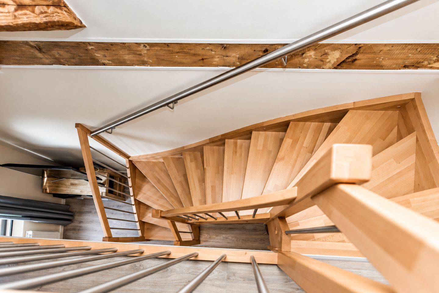 Una escalera de caracol de madera con barandillas de metal, integrada en un espacio con vigas de madera a la vista y techo blanco.