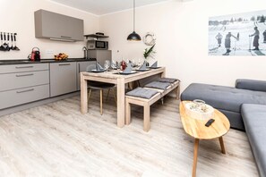 Une cuisine moderne avec des meubles gris, un four à micro-ondes et une bouilloire. On y trouve une table à manger avec des chaises, ainsi qu’une petite table ronde avec une télécommande. Un canapé gris et une étagère murale contenant des ustensiles de cuisine sont également visibles.