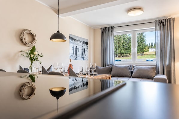 A modern living room with a dining area, featuring a sofa, a coffee table, and a dining table set with glasses and a bottle.