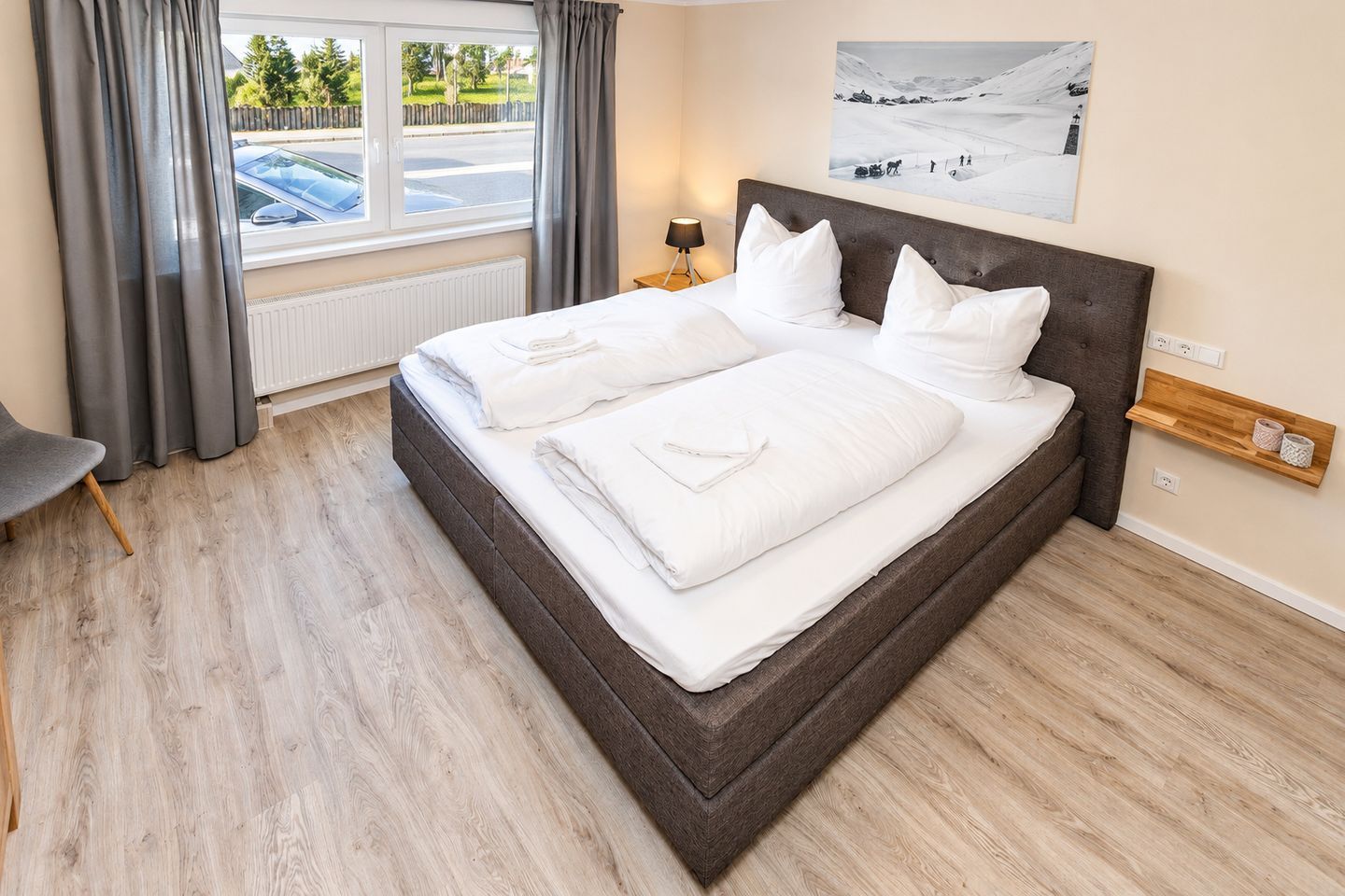 A neatly made bed with white linens in a room with light wooden flooring, a window with curtains, and a wall-mounted shelf.