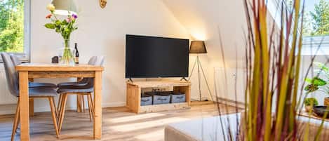 A modern living room with a wooden dining table, two chairs, a flat-screen TV on a wooden stand, a floor lamp, and a wall-mounted deer head decoration.