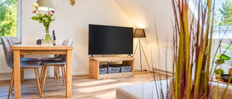 Ein modernes Wohnzimmer mit einem Holzesstisch, zwei Stühlen, einem Flachbildfernseher auf einem Holzständer, einer Stehlampe und einem an der Wand befestigten Hirsch-Dekoration.