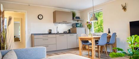 A modern kitchen with white cabinets, a dining area with a wooden table and chairs, a sofa, and a large window with greenery outside.
