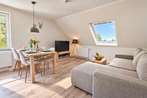 A modern living room with a dining area, a flat-screen TV, and a large window providing natural light. - Smart Resorts - Haus Opal - Ferienwohnung 404 mit 1 Schlafzimmer, Max. 4 Personen - Smart Resorts (Winterberg)