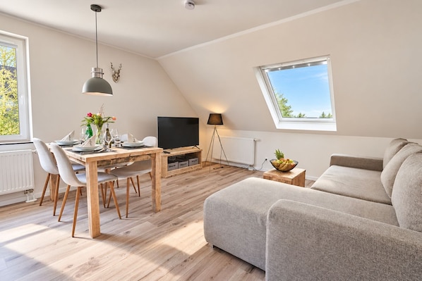 A modern living room with a dining area, a flat-screen TV, and a large window providing natural light.