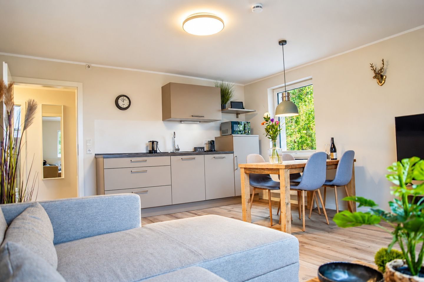 A modern kitchen with white cabinets, a dining area with a wooden table and chairs, a sofa, and a large window with greenery outside.