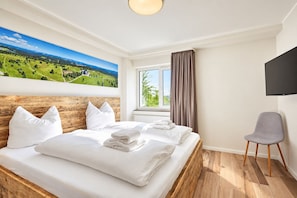 Habitación de hotel con una cama grande, cabecero de madera, vista a un paisaje verde por la ventana, un televisor de pantalla plana y una silla gris.