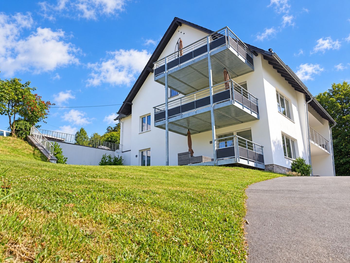 Ein zweistöckiges Gebäude mit einem Balkon im oberen Geschoss, einer Treppe zum Eingang und einer Grünfläche im Vordergrund.