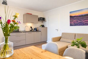 Een moderne keuken met een magnetron, een koffieapparaat en een theeset. Er is een eettafel met een bloemenvaas. Een bank staat in de woonkamer, met een schilderij aan de muur.