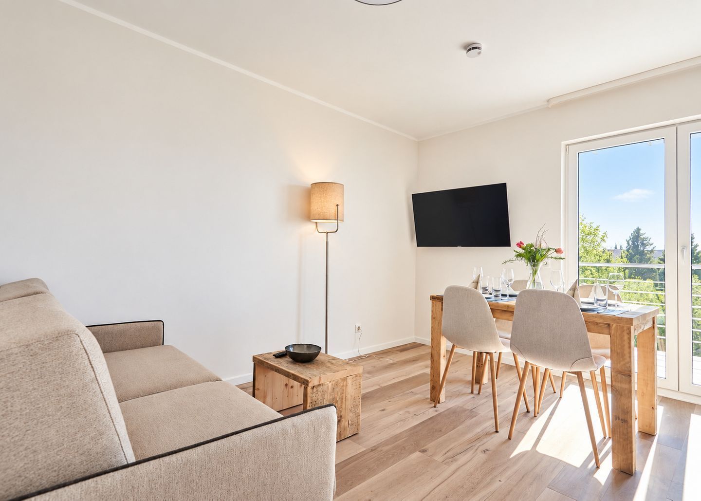 Una sala de estar moderna con un sofá, una mesa de comedor con sillas, un televisor de pantalla plana montado en la pared y un amplio ventanal con vistas a zonas verdes.