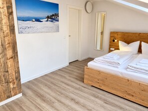 A bedroom with a wooden bed, white bedding, a large window, and a mountain landscape painting on the wall.