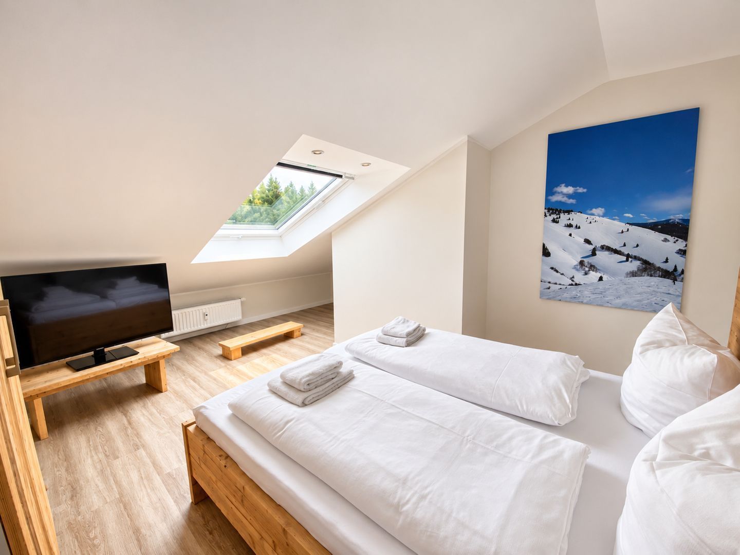 A bedroom with a TV, wooden bed, white bedding, a skylight, and a mountain landscape painting.