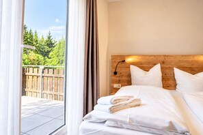 A hotel room with a wooden headboard, a bed with white linens, a nightstand with a lamp, and a view of a wooden deck and greenery outside.