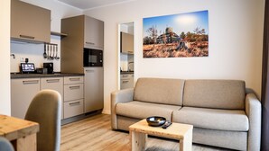 Een moderne woonkamer met een bank en salontafel, een keuken met roestvrijstalen apparatuur, en een schilderij van een landschap aan de muur.