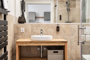 A bathroom with a wooden vanity, a white sink, a mirror, a towel rack, a soap dispenser, and a shower area.