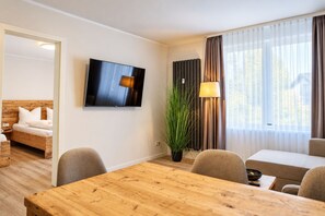 A modern hotel room with a wooden dining table, a sofa, a TV mounted on the wall, and a large window with curtains.