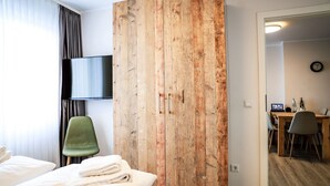 Une chambre d’hôtel avec une grande armoire en bois, un lit avec du linge de maison blanc, une chaise verte et un bureau avec un ordinateur et une bouteille.