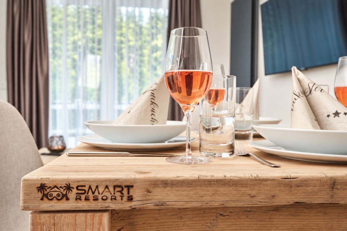 Una mesa de comedor con una copa de vino rosado, platos y una servilleta con estampado de estrellas. La mesa está sobre una superficie de madera con un logotipo que dice 'SMART RESORTS'.