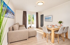 A modern living room with a sofa, dining table, and chairs. There is a painting on the wall and a sign that reads 'SMART RESORTS'. - Smart Resorts - Haus Azur - Ferienwohnung 805, Max. 5 Personen - Smart Resorts Haus Azur (Winterberg)