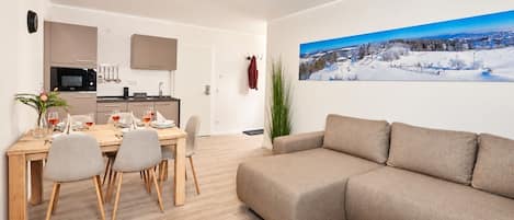 A modern living room with a sectional sofa, a dining area with a wooden table and chairs, a kitchen with a microwave and a TV, and a large window with a snowy landscape view.
