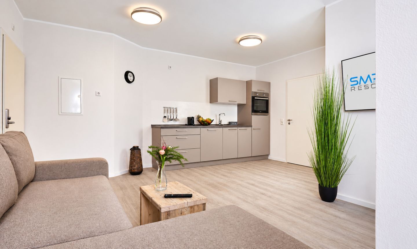 A modern living room with a sofa, a small wooden table with a vase, and a kitchen area in the background.