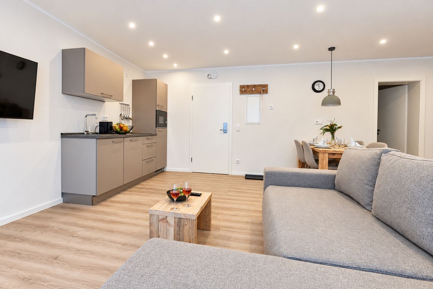 A modern living room with a grey sofa, a wooden coffee table with glasses, and a kitchen area with cabinets and a TV.