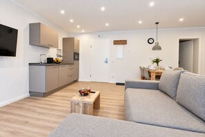 A modern living room with a grey sofa, a wooden coffee table with glasses, and a kitchen area with cabinets and a TV.