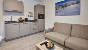 Ein modernes Wohnzimmer mit einer Couch, einem Couchtisch mit Früchten, einer Küche mit einer Mikrowelle und einem Gemälde einer verschneiten Landschaft.