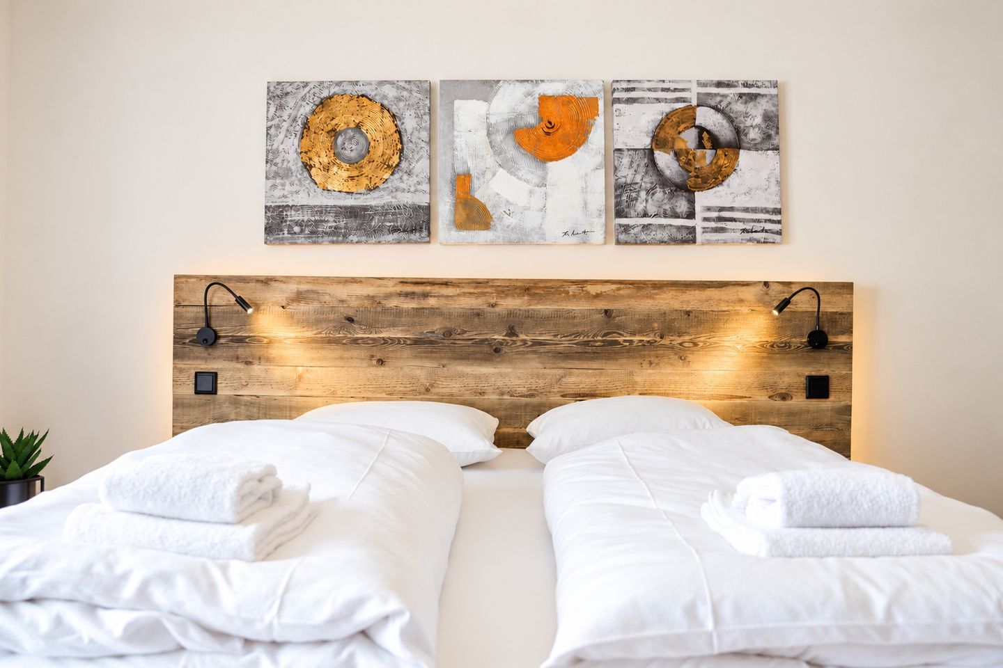 Una habitación de hotel con cabecera de madera, dos camas con sábanas blancas y tres cuadros abstractos sobre ellas.