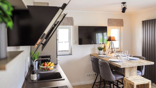 A modern kitchen with a built-in oven, a dining area with a wooden table and chairs, a flat-screen TV mounted on the wall, and a window providing natural light.