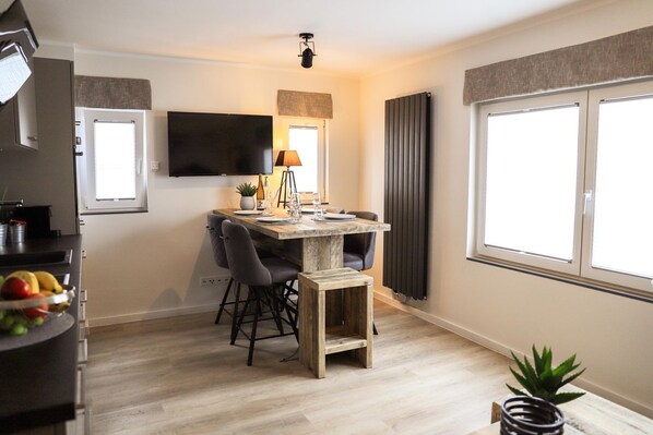 A compact living space with a dining area, a flat-screen TV mounted on the wall, a window with curtains, and a radiator.