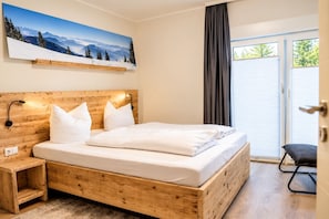 Un dormitorio con cama de madera, ropa de cama blanca, una mesita de noche, una lámpara de pared y un amplio ventanal con persianas.