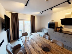 Un salon moderne avec une table à manger en bois, un canapé, un coin cuisine et un téléviseur fixé au mur.