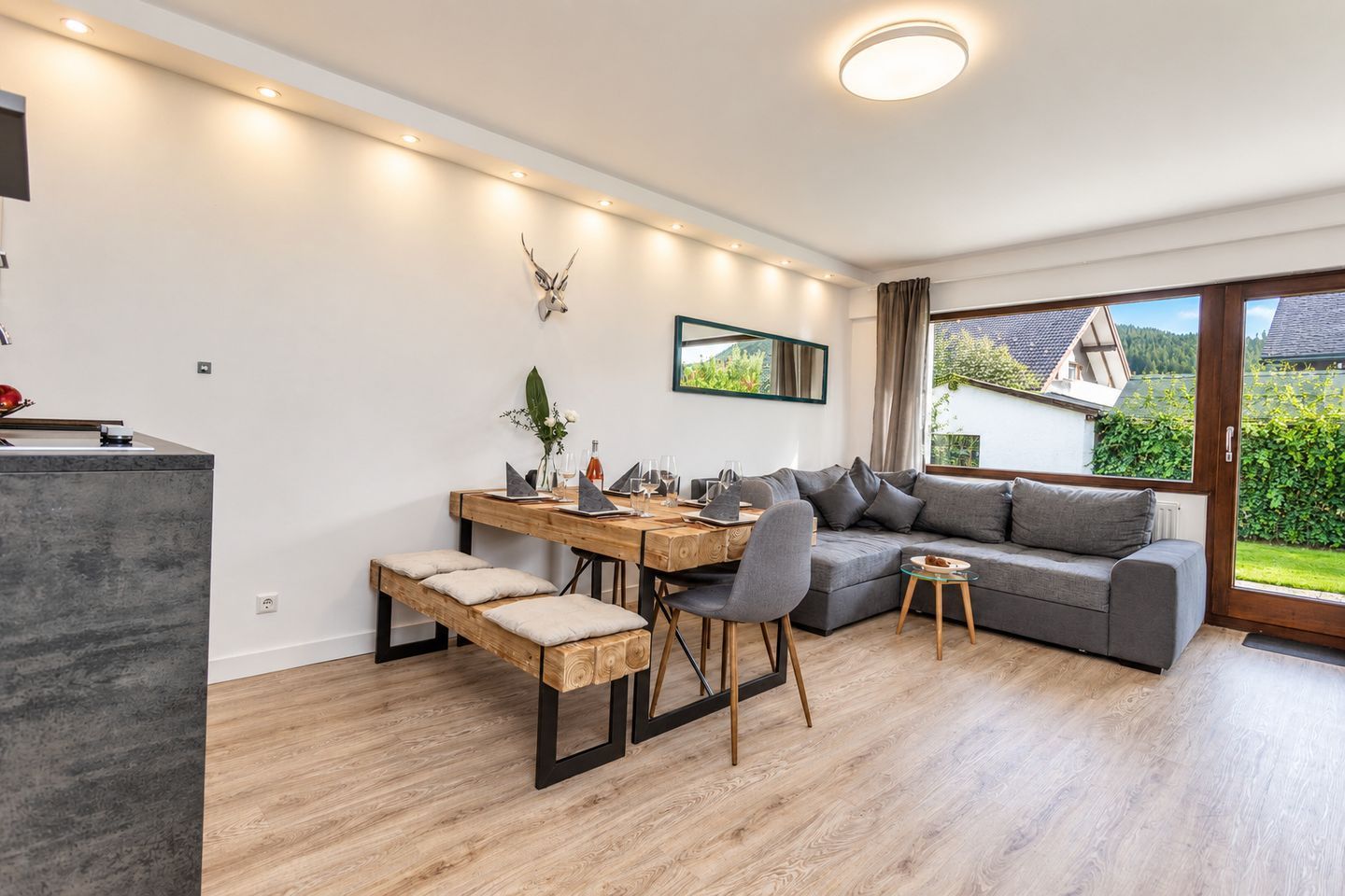 A modern living space with a dining area, a sofa, and a large window offering a view of the outdoors.