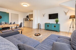 A modern living room with a sofa, a dining table with chairs, a kitchen area, and a mounted television.
