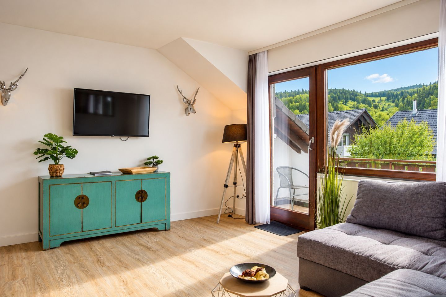 A modern living room with a teal cabinet, a flat-screen TV, a tripod lamp, and a view of a forested landscape through a large window.