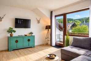 A modern living room with a teal cabinet, a flat-screen TV, a tripod lamp, and a view of a forested landscape through a large window.
