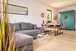 Una sala de estar moderna con un sofá gris, una mesa de comedor con sillas y una zona de cocina con un armario verde azulado.