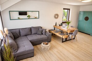 A modern living room with a grey sectional sofa, a wooden dining table with chairs, a small side table, and a large mirror on the wall. - Smart Resorts - Haus Rubin - Ferienwohnung 107 mit 2 Schlafzimmern und Balkon, Max. 6 Personen - (Winterberg)