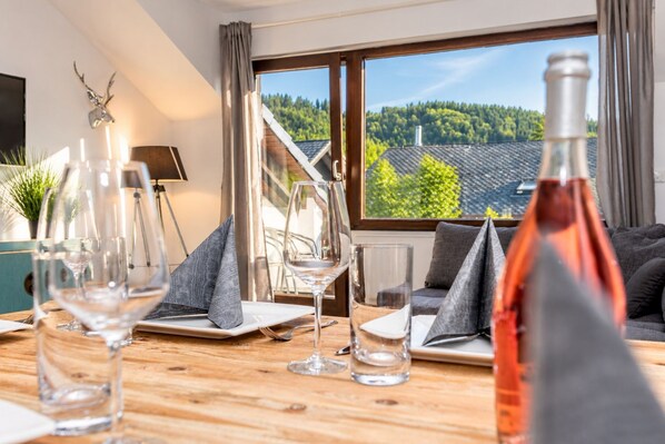 A dining area with a wooden table set for two, a bottle of red wine, and a view of a forest through large windows.