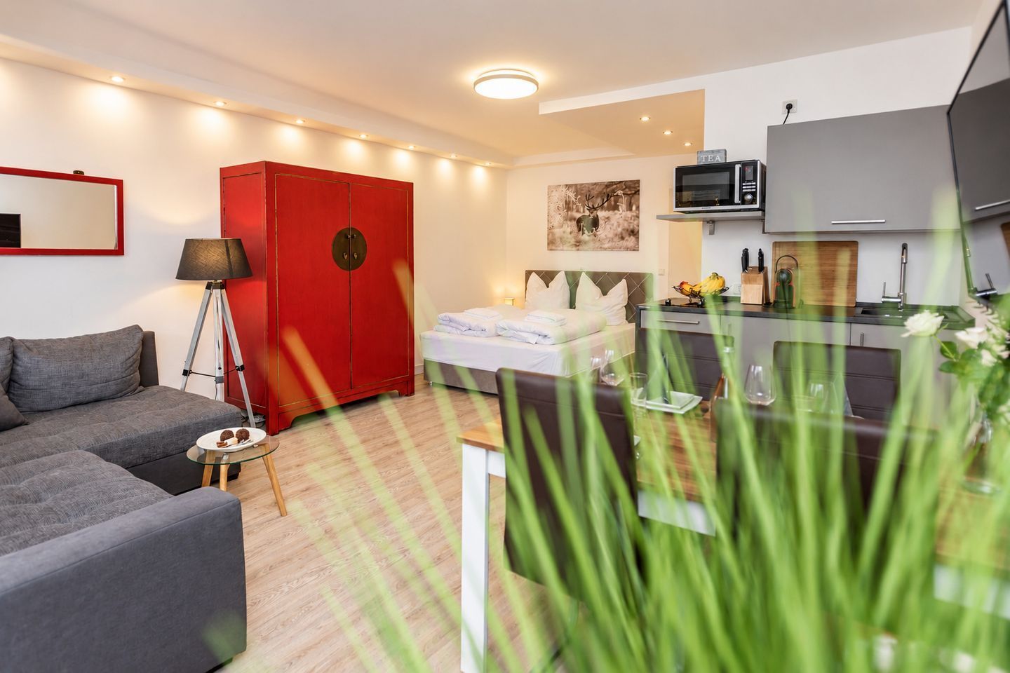 A modern hotel room with a bed, a red wardrobe, a sofa, a desk with a chair, and a kitchenette.