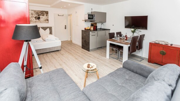 Ein modernes Wohnzimmer mit grauen Sofas, einem kleinen runden Tisch mit Snacks, einem roten Schrank, einer Küchenzeile mit Mikrowelle und einem Esstisch für zwei Personen.