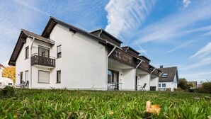 Ein zweigeschossiges Wohngebäude mit Balkon, überdachter Terrasse und Rasenfläche vor der Fassade.