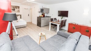 Ein modernes Wohnzimmer mit einem grauen Sofa, einem kleinen runden Tisch mit Snacks, einem roten Schrank und einer Küchenzeile mit Mikrowelle und Fernseher.