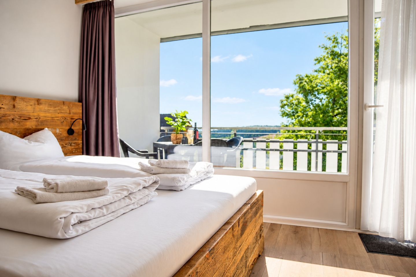 A hotel room with a large bed, a wooden headboard, a balcony with a view of greenery and a body of water, and a dining area with a table and chairs.