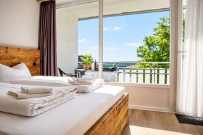 A hotel room with a large bed, a wooden headboard, a balcony with a view of greenery and a body of water, and a dining area with a table and chairs.